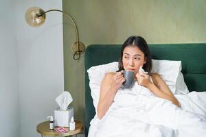 santé et personnes. asiatique fille mensonge dans lit, sentiment malade, éternuements et en buvant chaud thé, contagieux froid, rester à Accueil sur laisser photo