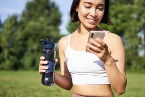 souriant aptitude fille les boissons eau, chèques sa app sur téléphone intelligent et regards content, séjours hydraté sur Frais air, ensoleillé journée dans parc photo