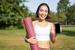 souriant asiatique aptitude fille avec caoutchouc yoga tapis, spectacles sa téléphone intelligent filtrer, recommande faire des exercices application, des stands sur pelouse dans parc photo