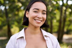 gens concept. souriant asiatique fille à la recherche à caméra avec content, joyeux émotion, posant dans vert parc sur une chaud été journée photo