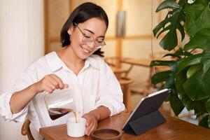 free-lance et éloigné ouvriers. souriant Jeune femme verser café dans une tasse, séance dans café et à la recherche à numérique tablette photo