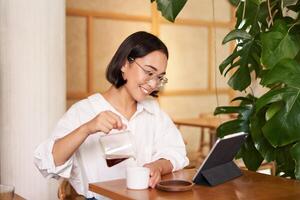 free-lance et éloigné ouvriers. souriant Jeune femme verser café dans une tasse, séance dans café et à la recherche à numérique tablette photo