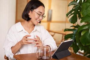 asiatique femme dans lunettes, en train de regarder qch sur numérique tablette, en buvant café dans une café et souriant, travail à distance photo