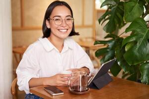 souriant asiatique fille dans lunettes, femme travail sur télécommande, en buvant café et en utilisant tablette photo