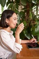magnifique asiatique fille séance dans café avec tasse de café, dessin sur numérique tablette avec graphique stylo, gribouiller et fabrication les taches photo