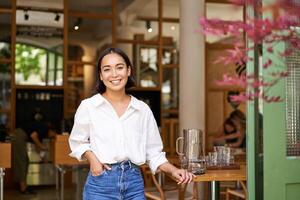 portrait de Jeune femme d'affaires dans sa posséder café, directeur permanent près entrée et attrayant toi, posant dans blanc plaine chemise et jeans photo