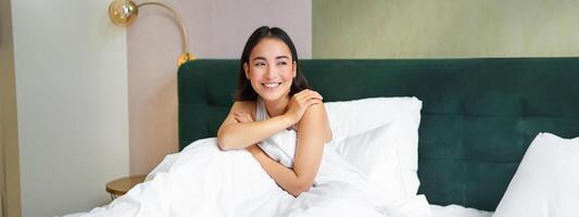 content les matins. souriant asiatique femme se réveille en haut dans sa lit, regards à l'extérieur et ressenti enthousiaste à propos sa Matin. chambre intérieur concept photo