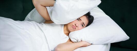 triste fille avec insomnie. Jeune asiatique femme mensonge dans lit, couverture sa oreilles avec oreiller, cant dormir, partenaire ronfle, bruyant bruit à nuit inquiétant sa photo