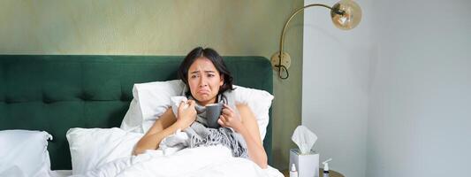 malade triste asiatique fille mensonge dans sa lit avec grippe, capture une froid, prise pilules et en buvant chaud thé, sentiment dérangé photo