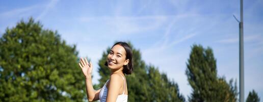 portrait de asiatique fille sur aptitude classe dans parc, séance sur caoutchouc tapis et vague main à caméra, dire Bonjour, méditer et entraine toi yoga photo