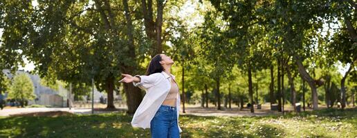 insouciant asiatique fille dansant, sentiment bonheur et joie, profiter le Soleil sur été jour, en marchant dans parc avec vert des arbres photo