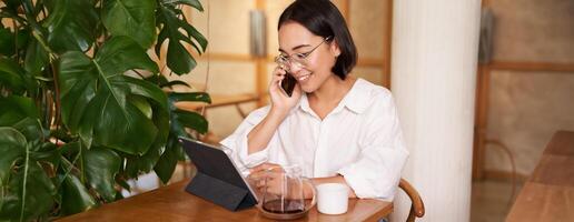 femme dans lunettes, répondre téléphone appel dans une café, en buvant café, à la recherche à travail sur tablette, travail sur éloigné de restaurant photo