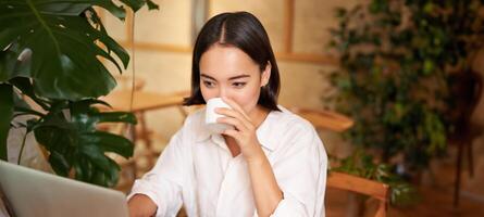 étudiant séance dans café avec tasse de café. Jeune asiatique femme travail sur portable dans restaurant, séance avec ordinateur et téléphone intelligent photo