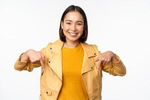 portrait de content souriant asiatique fille, montrer du doigt les doigts vers le bas et montrant logo, démontrant bannière, permanent dans Jaune veste contre blanc Contexte photo