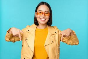 portrait de élégant asiatique fille, porte des lunettes de soleil, sourit et points les doigts bas, spectacles publicité, des stands plus de bleu Contexte photo