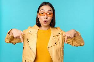 portrait de élégant asiatique fille, porte des lunettes de soleil, sourit et points les doigts bas, spectacles publicité, des stands plus de bleu Contexte photo