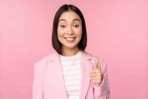 portrait de asiatique femme d'affaires souriant satisfait, montrant les pouces en haut, louer, comme et approuver, permanent dans costume plus de rose Contexte photo