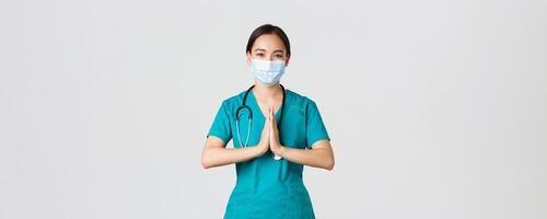 COVID-19 [feminine, coronavirus maladie, soins de santé ouvriers concept. optimiste souriant asiatique femelle médecin, médecin dans médical masque et gommages tenir mains dans prier geste, remerciement, blanc Contexte photo