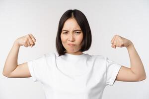 souriant asiatique femme montrant fléchissement biceps, muscles fort bras geste, permanent dans blanc T-shirt plus de blanc Contexte photo