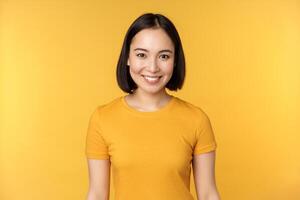portrait de Jeune moderne asiatique femme, souriant content avec blanc dents, à la recherche sur de soi à caméra, portant décontractée T-shirt, permanent plus de Jaune Contexte photo