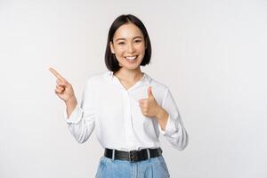 image de Jeune asiatique affaires femme, souriant tandis que montrer du doigt doigt la gauche et montrant les pouces en haut, recommander produit, louer, permanent plus de blanc Contexte photo