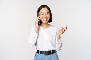 souriant content asiatique femme parlant sur téléphone intelligent avec client, vendeuse sur appel, en portant mobile téléphone et faire des gestes, permanent plus de blanc Contexte photo