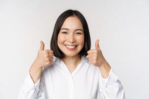 de bonne humeur asiatique femme affronter, montrant les pouces en haut et souriant, heureux par quelque chose, recommander qch bien, permanent plus de blanc Contexte photo