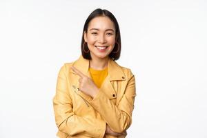 portrait de souriant asiatique fille montrer du doigt doigt gauche, montrant logo ou copie espace, publicité sur vide côté, permanent plus de blanc Contexte photo