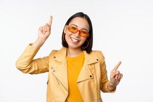 souriant asiatique brunette femme dans des lunettes de soleil, montrer du doigt les doigts de côté, la gauche et droite, montrant variantes, en riant et dansant, portant des lunettes de soleil, blanc Contexte photo