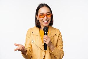 portrait de magnifique asiatique femme dans des lunettes de soleil, élégant fille en chantant, donnant discours avec microphone, en portant micro et souriant, permanent plus de blanc Contexte photo