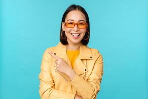 souriant asiatique femme dans des lunettes de soleil, montrer du doigt doigt gauche, montrant bannière, logo ou publicité, permanent plus de bleu Contexte photo