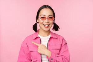 portrait de souriant asiatique fille dans élégant tenue, des lunettes de soleil, montrer du doigt doigt gauche, montrant publicité, bannière, permanent plus de rose Contexte photo