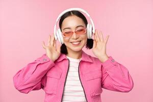 dansant élégant asiatique fille écoute la musique dans écouteurs, posant contre rose Contexte photo