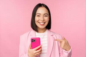 portrait de souriant asiatique femme d'affaires montrer du doigt à sa mobile téléphone, recommander téléphone intelligent application, application sur téléphone portable, permanent plus de rose Contexte photo