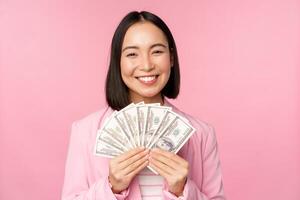 finance, microcrédit et gens concept. content souriant asiatique femme d'affaires montrant dollars argent, permanent dans costume contre rose Contexte photo