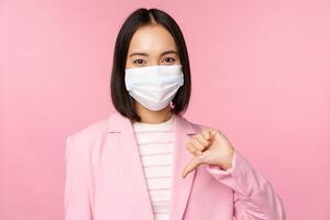 coréen affaires femme dans médical visage masque et costume, spectacles les pouces bas, Ne pas aimer ou désapprouver geste, permanent plus de rose Contexte photo