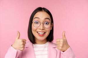 génial, bravo. visage de excité asiatique femme d'affaires dans lunettes, souriant heureux, montrant les pouces en haut dans approbation, permanent plus de rose Contexte photo