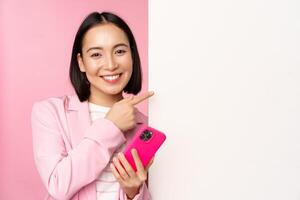 image de souriant asiatique entreprise femme dans costume, en portant téléphone intelligent, montrer du doigt à conseil, montrant graphique ou information logo sur vide blanc mur, permanent plus de rose Contexte photo