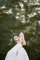 une fille dans lumière rayé un pantalon accroché sa jambes dans des sandales plus de le l'eau surface. photo