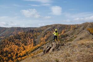 une gars dans brillant tenue de sport des promenades le long de le Haut de le montagne, regards vers l'avant à le montagnes et forêt, clair ciel aérien, l'automne paysage photo