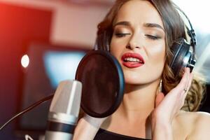 beau chanteur avec écouteurs dans de face de microphone chante avec sa ouvert bouche dans moderne studio. agréable femme dans écouteurs à le micro chante avec un expression de l'amour. photo