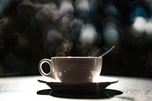 blanc tasse avec chaud liquide et vapeur sur noir flou Contexte. cuillère à café dans une Coupe. thé et café concept. photo