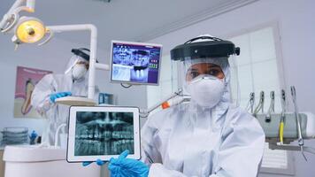 patient pov dans dentaire Bureau avec Nouveau Ordinaire discuter traitement de les dents cavité, dentiste montrer du doigt sur numérique radiographie en utilisant tablette. stomatologie portant protecteur Hazmat costume contre coroanvirus photo