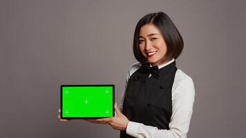 femme serveuse en portant tablette avec écran vert afficher sur caméra, en présentant Vide maquette modèle dans studio. asiatique restaurant employé montrant dispositif fonctionnement chromakey filtrer. caméra b. photo