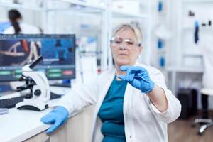 Sénior scientifique dans protecteur équipement en portant échantillon pendant clinique étude donnant compétence. personnes âgées chercheur dans stérile laboratoire à la recherche sur microscope faire glisser portant laboratoire manteau. photo