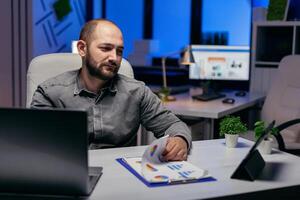 homme d'affaire travail en retard à nuit dans lieu de travail en utilisant tablette PC en portant main sur presse-papiers avec graphique. barbu entrepreneur Faire heures supplémentaires à le Bureau dans le soir à terminer une date limite. photo