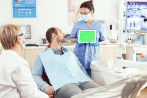 malade homme à la recherche à moquer en haut vert écran chrominance clé tablette avec isolé afficher tandis que séance à dentaire chaise dans hôpital stomatologie clinique bureau. médical équipe expliquant traitement mal aux dents photo
