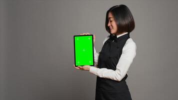 femme serveuse en portant tablette avec écran vert afficher sur caméra, en présentant Vide maquette modèle dans studio. asiatique restaurant employé montrant dispositif fonctionnement chromakey filtrer. caméra un. photo