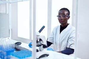 femme de africain l'ethnie travail dans soins de santé établissement séance à sa lieu de travail avec protecteur lunettes. noir soins de santé scientifique dans biochimie laboratoire portant stérile équipement. photo