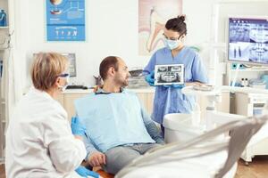 malade homme séance sur stomatologique chaise écoute médecin tandis que à la recherche à tablette dans dentaire clinique. dentisterie médecin montrant à patient les dents radiographie pendant dentaire consultation photo